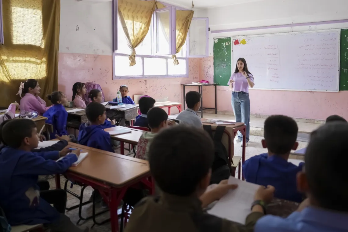A female teacher stands at the front of a classroom filled with children.