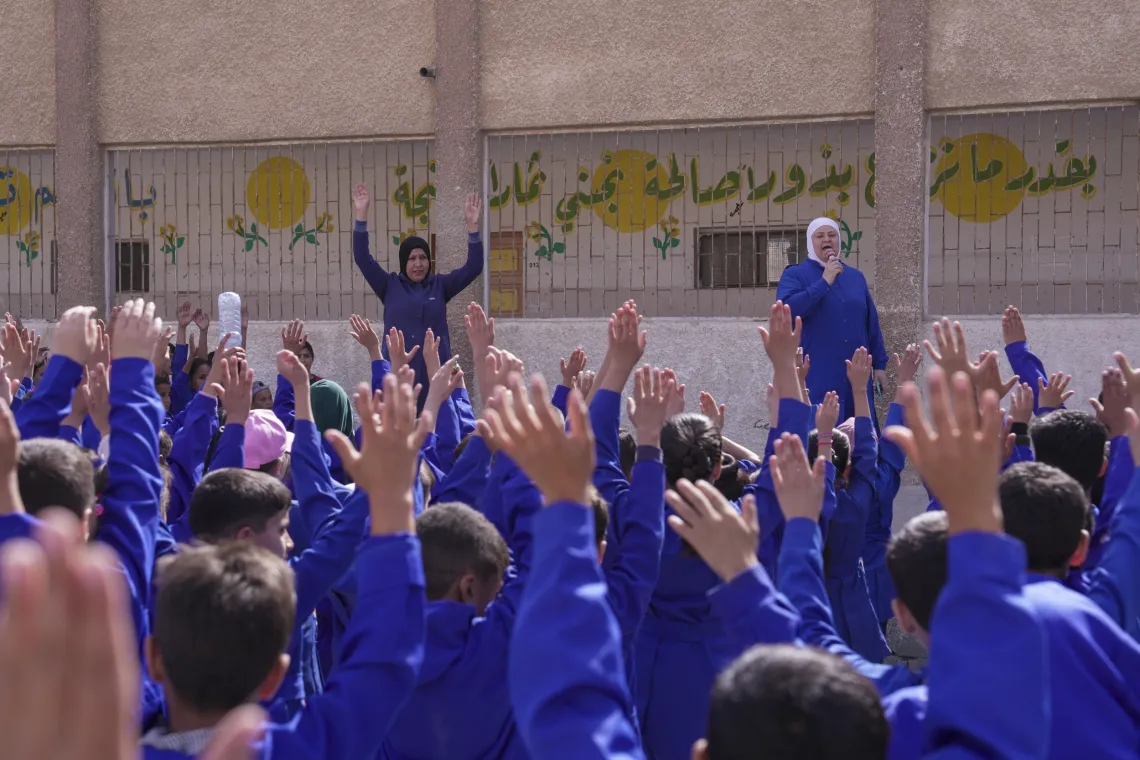 Looking from behind a large group of school children dressed in blue, some with their arms lifted into the air.