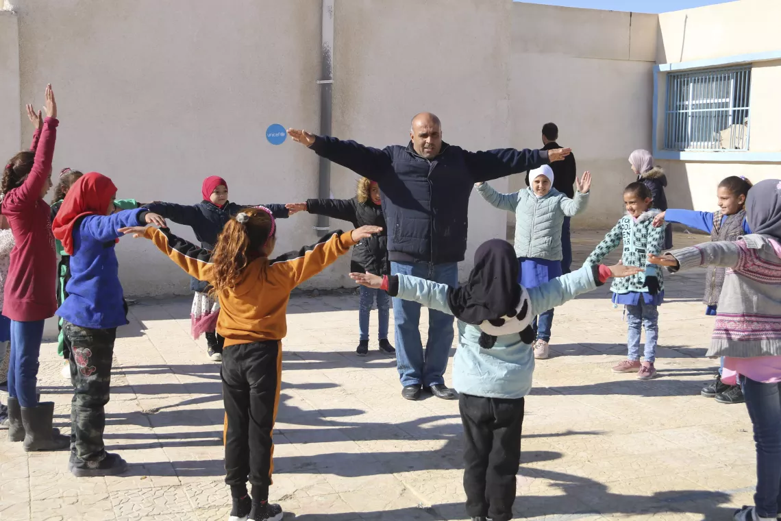A man stands with his arms stretched out on his sides, surrounded by a large circle of young children doing the same.