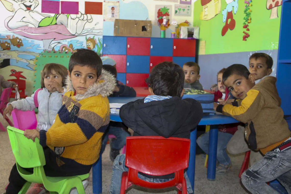 Small children sitting on colourful chairs in a brightly decorated classroom.