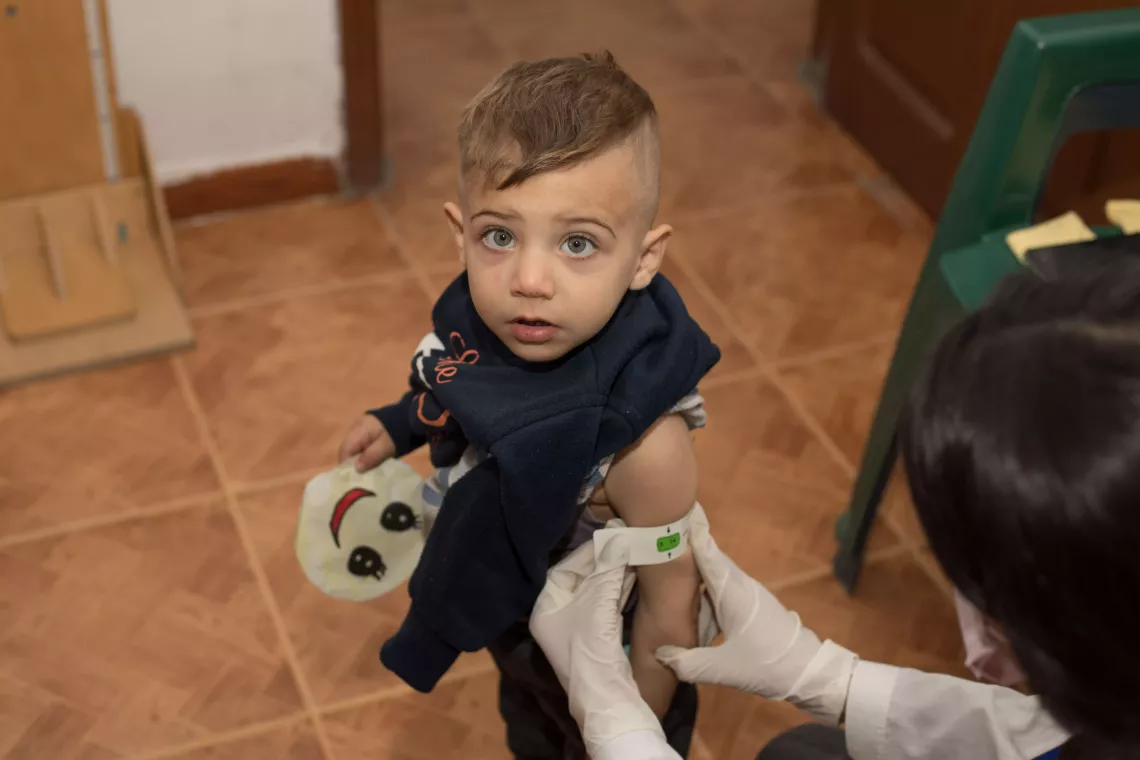 A little boy looks up at the camera with bright eyes, while a nutrition worker measures his arm circumference. 