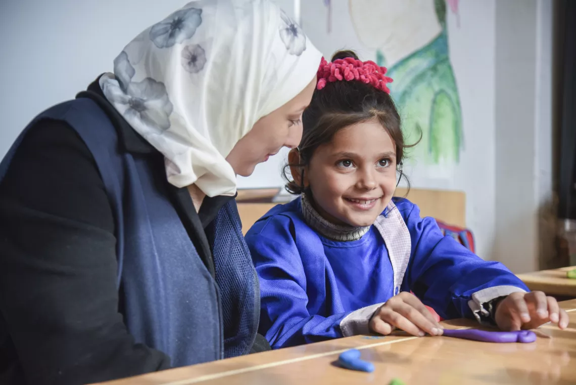 A little girl smiles while looking off into the distance while the woman sitting next to her is talking to her.