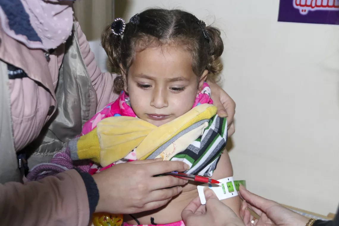 A little girl with hair tied in pigtails and her sleeve pulled up, looks down while her mid arm circumference is measured.