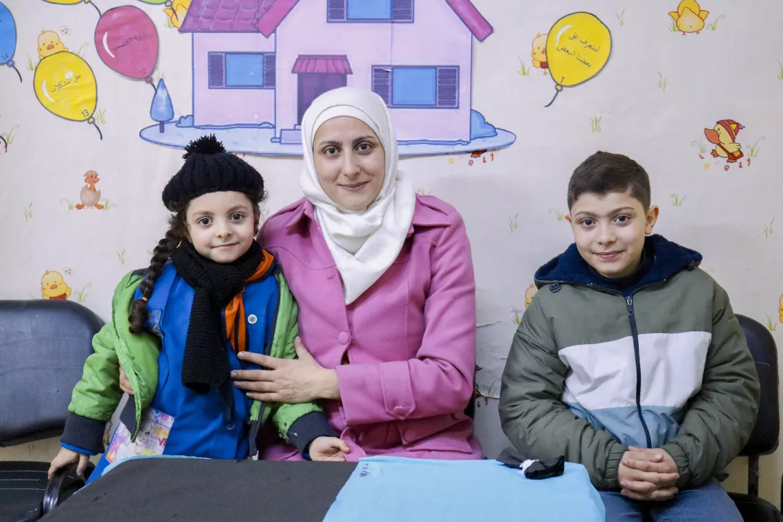 A mother wearing a pink coat and white headscarf sits between her son and daughter, all smiling brightly.