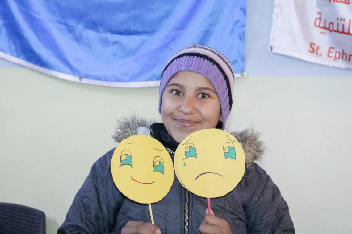 A wearing a warm hat smiles while holding up a happy and a sad face drawn on rounds of paper.