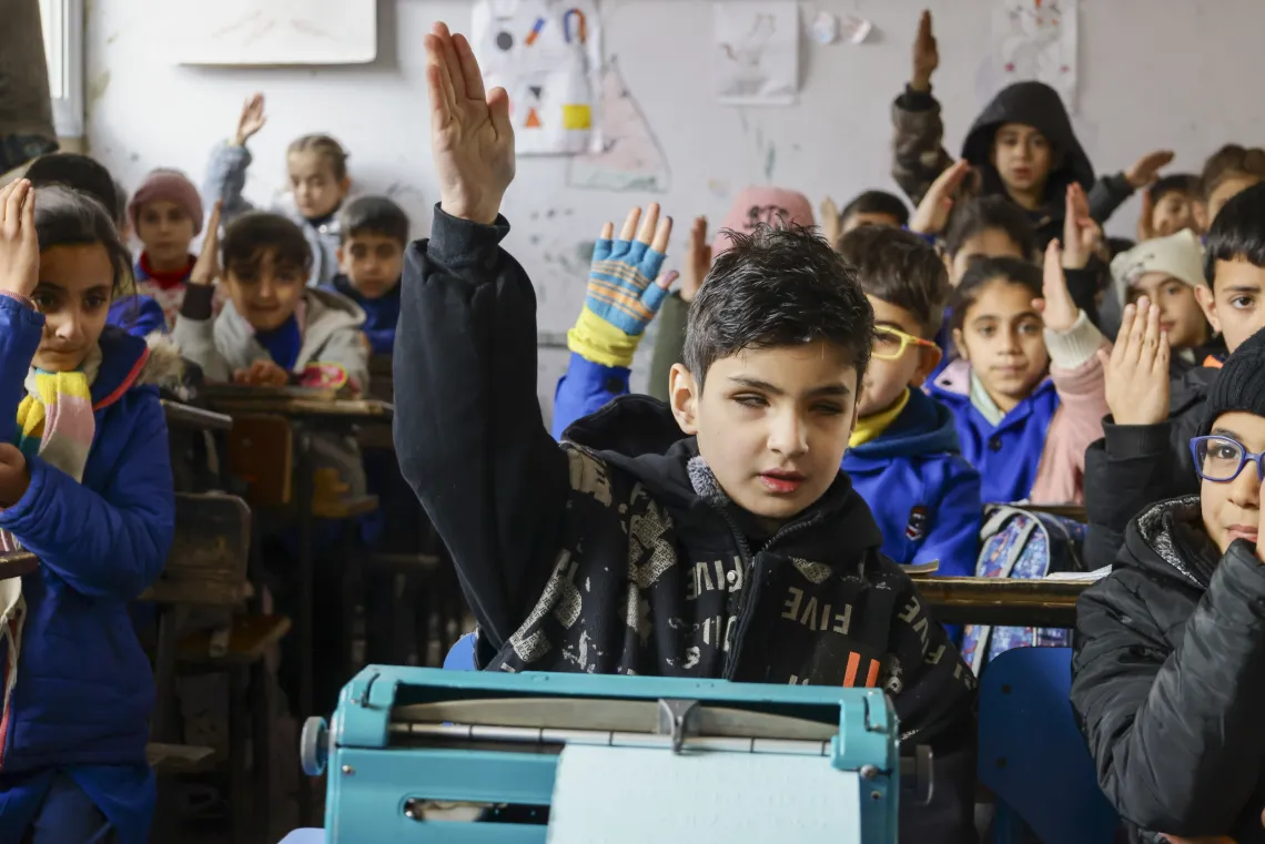 Youssef has his hand up to answer a question in class. He sits at a desk in the front of the class with his braille machine on the desk in front of him.