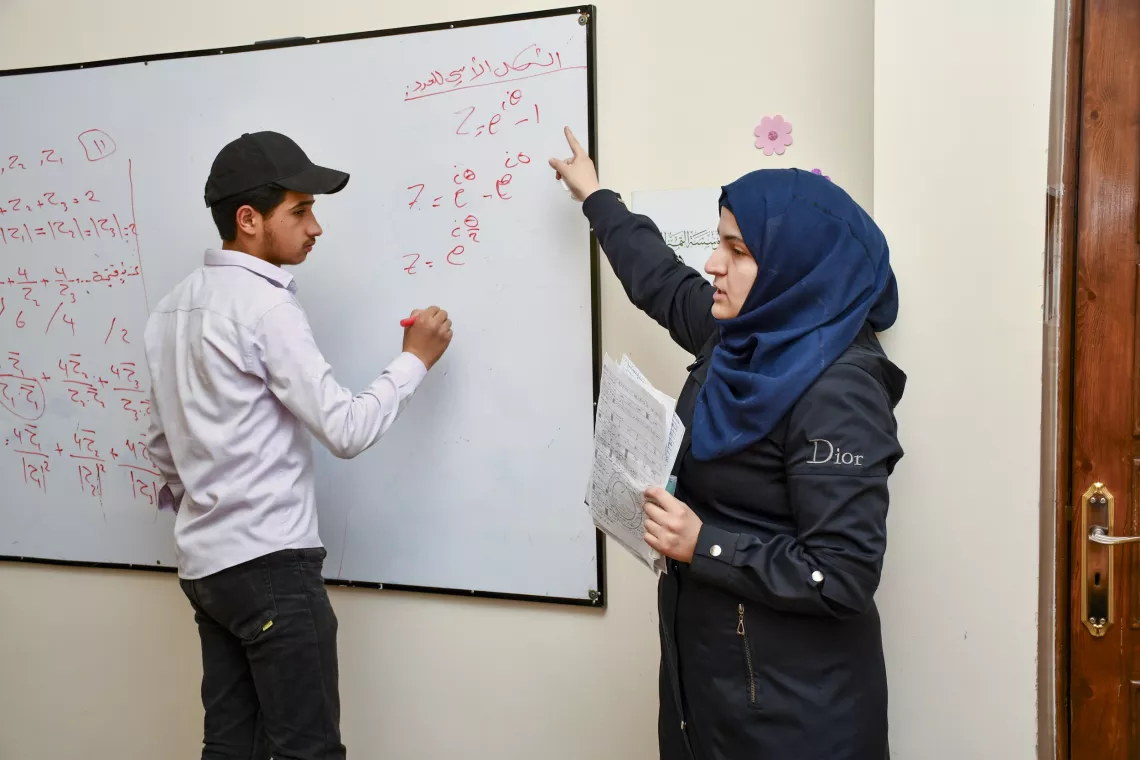 Suleiman, 19, engaged in learning, while writing on the whiteboard as Ms. Rama, the teacher, guides him.