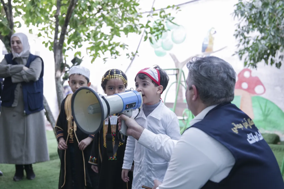A little boy speaks on a megaphone that is held by a health worker.