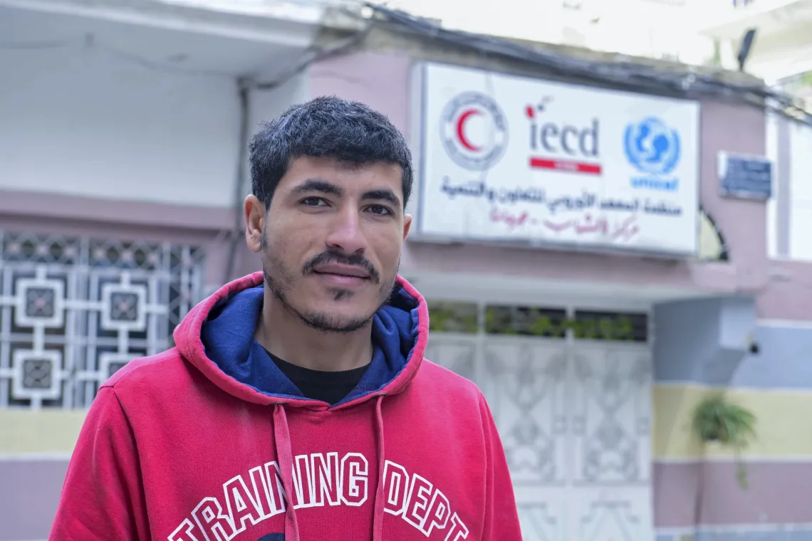 Portrait of a young man wearing a red hoodie outside a building with a UNICEF and other logos on it.
