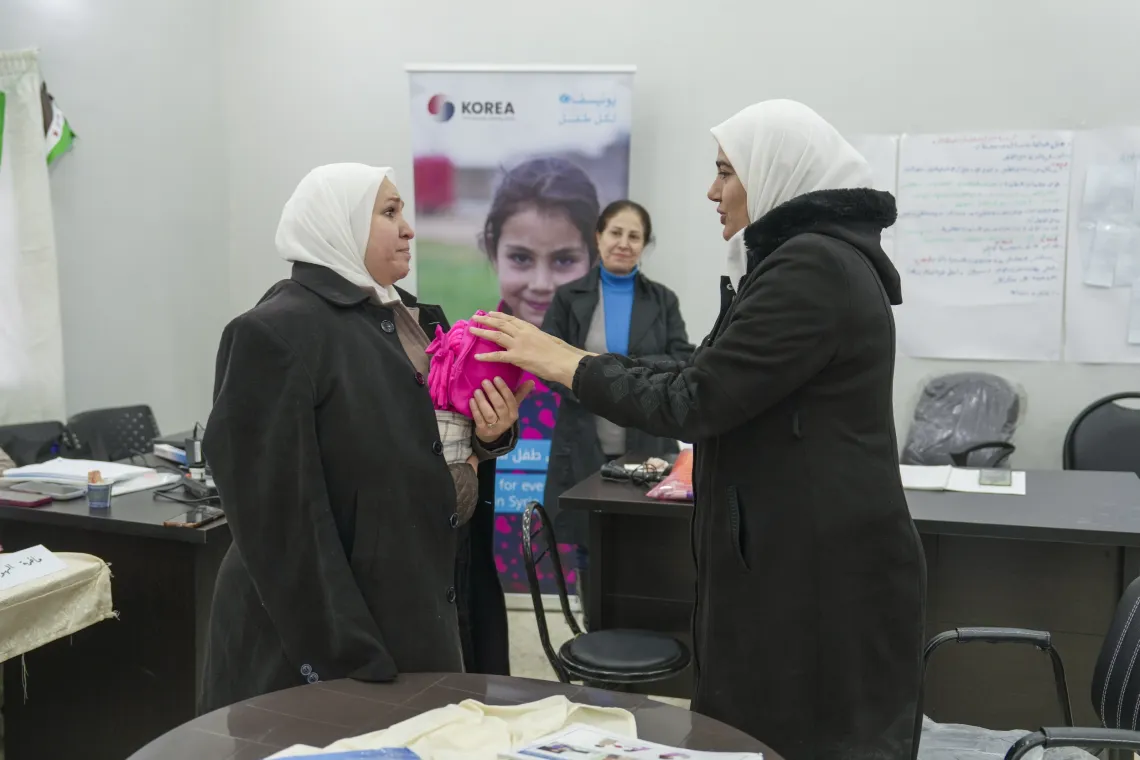A woman holds a pretend infant to anothe woman's chest, while explaining something to her.