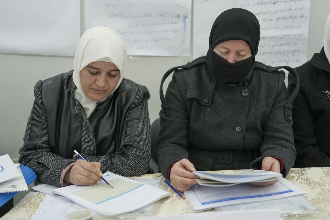 Two women sitting at a desk looks down at their papers, while listening and taking notes.