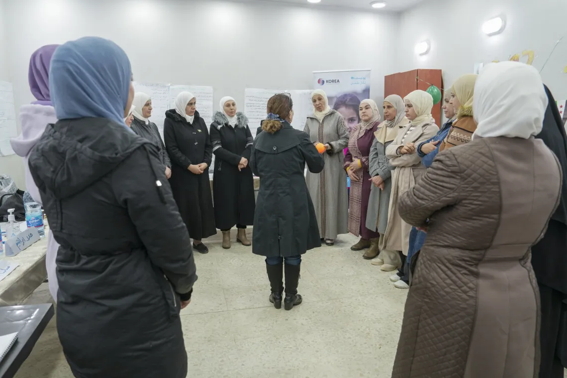 A group of women are standing in a big circle, listening to a woman speaking who is standing in the middle.