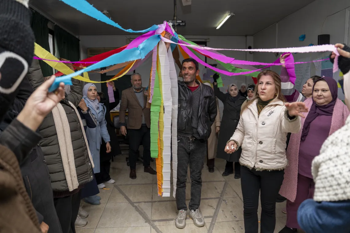 A group of people stand in a circle each holding a coloured ribbon tied together in the middle of the group, with a woman explaining something to them.