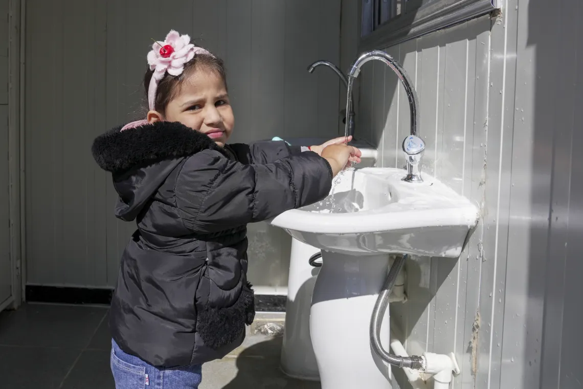 A little girl with a headband with a pink flower on it, washes here hands at a sink.
