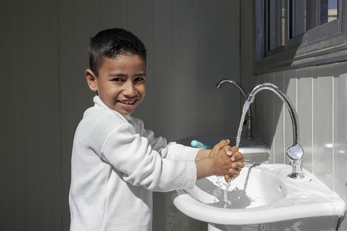 A young boy smiles as he washes his hands in a sink.