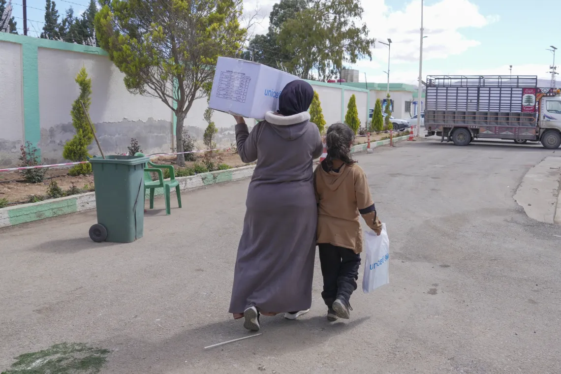 A woman carrying a large box on her shoulder, walks next to her daughter who is carrying a while bag.