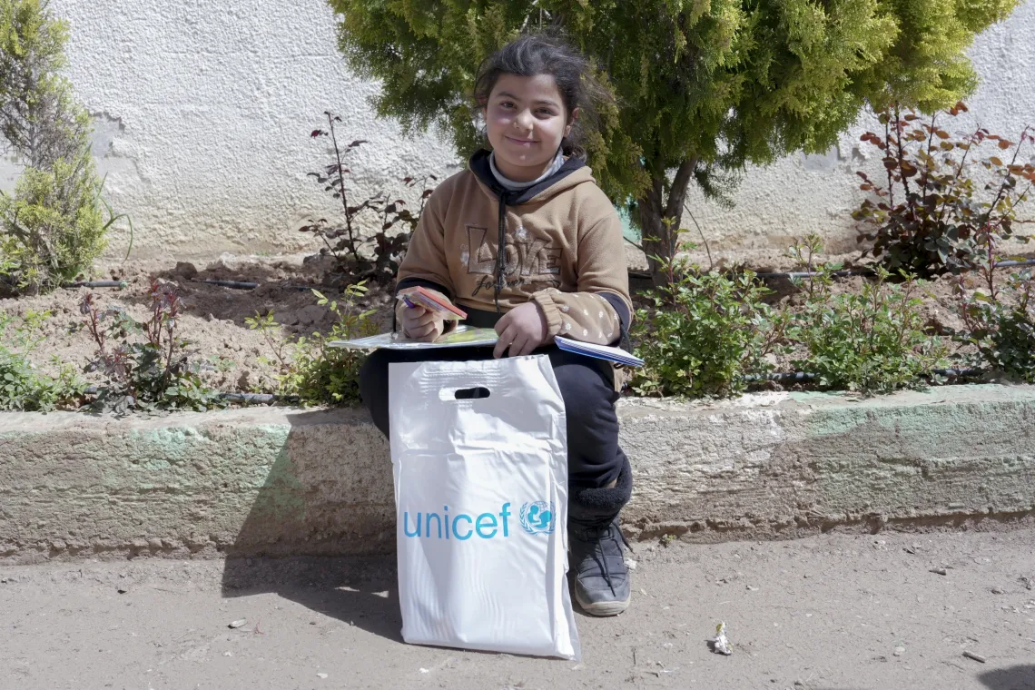 A little girl sits on the curbside with a white bag with a UNICEF logo on it in front of her.