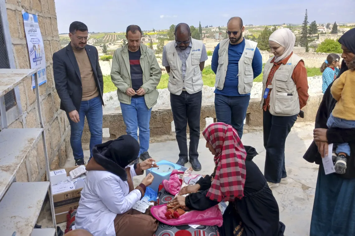 A vaccinator prepares injection to provide vaccine to a child with UNICEF and government staff looking on.
