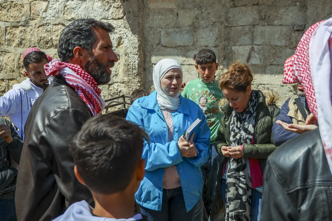 A woman wearing a blue UNICEF coat speaks members of the community who are standing around her.