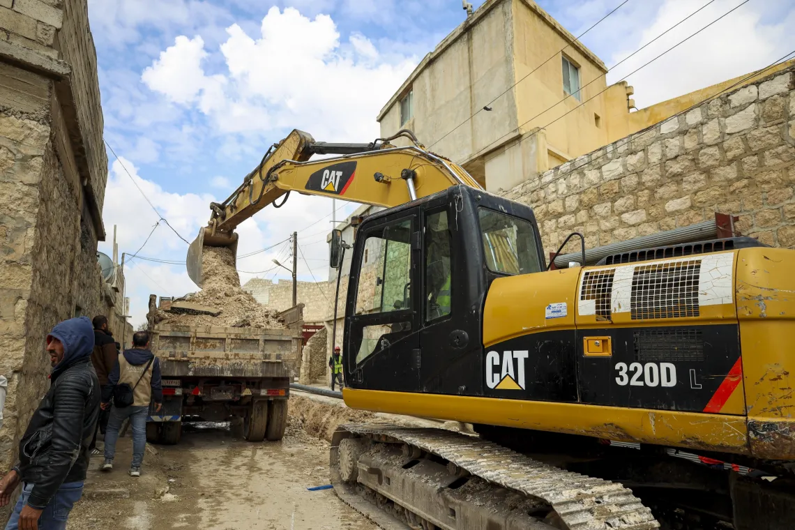 A large yellow excavator in a narrow street loads dirt on a waiting truck.