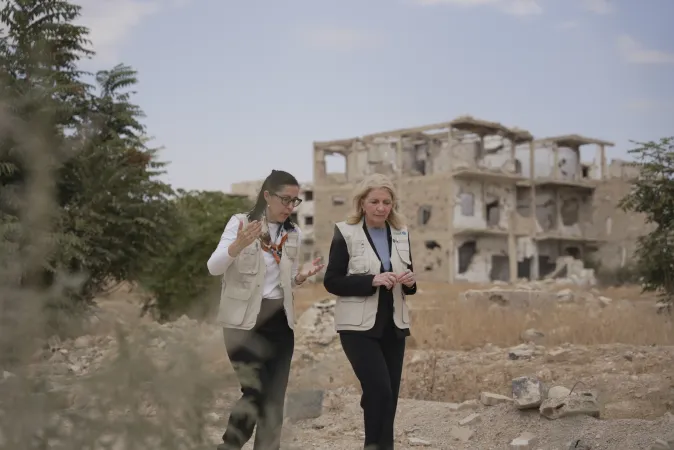 Two women walk in a dusty path with the ruins of a building in the background