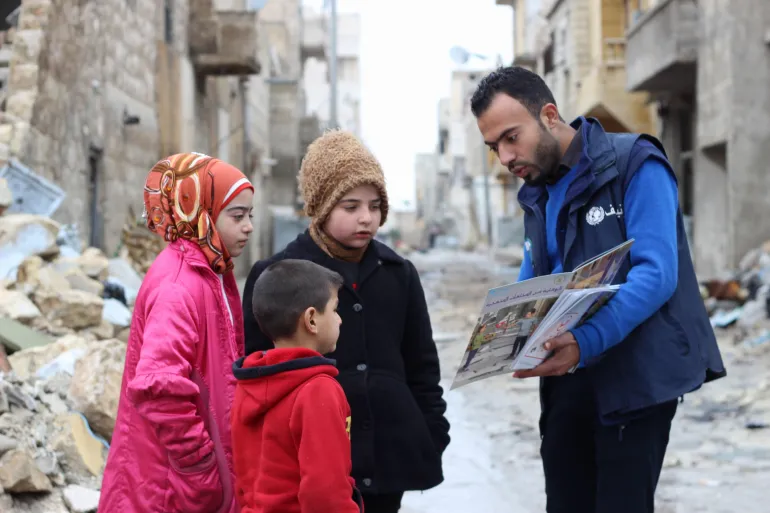 A volunteer show pictures to three young children in a street with rubble and war damaged buildings.