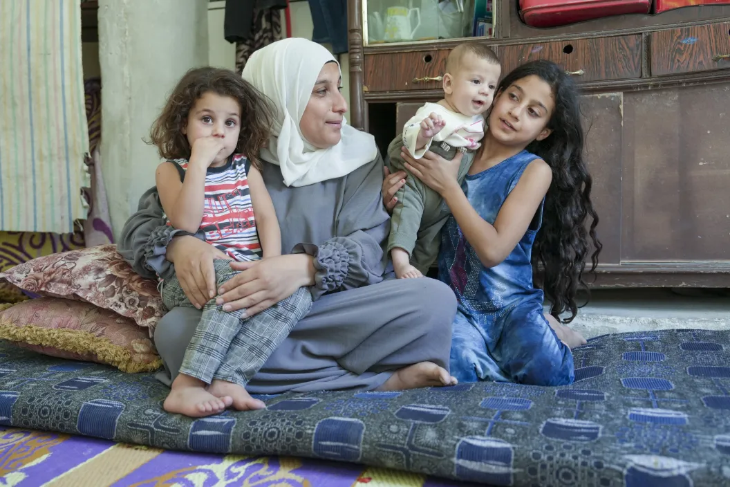 A woman, two girls and a little baby sit on a carpet. One girl is sitting partially on her mom's lap, held by her mom, while her sister is holding her baby brother.