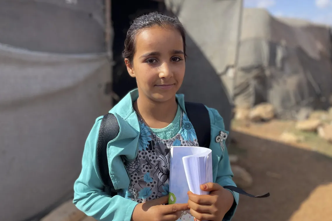 A girl standing outside a tented shelter holds a book in her hands.