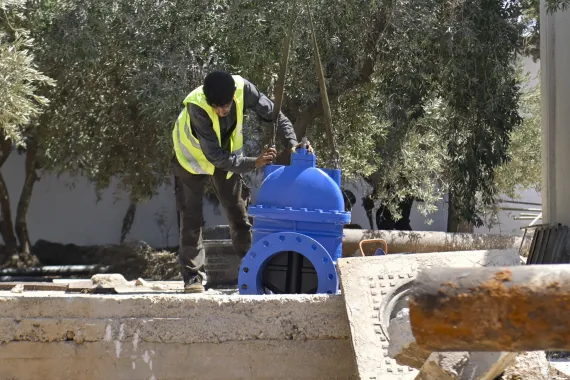 A worker wearing a yellow reflective vest bends down to inspect a large blue valve.