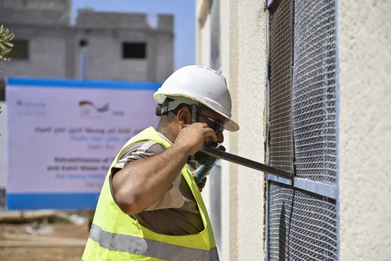 A man wearing a white hardhat and yellow reflective vest uses a large tool to install a metal mesh.