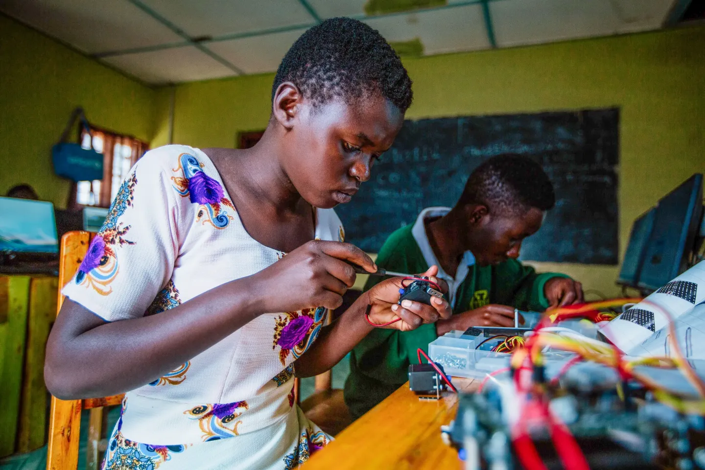 Student soldering wires in a classroom workshop, focused, with another student assembling electronics in the background.