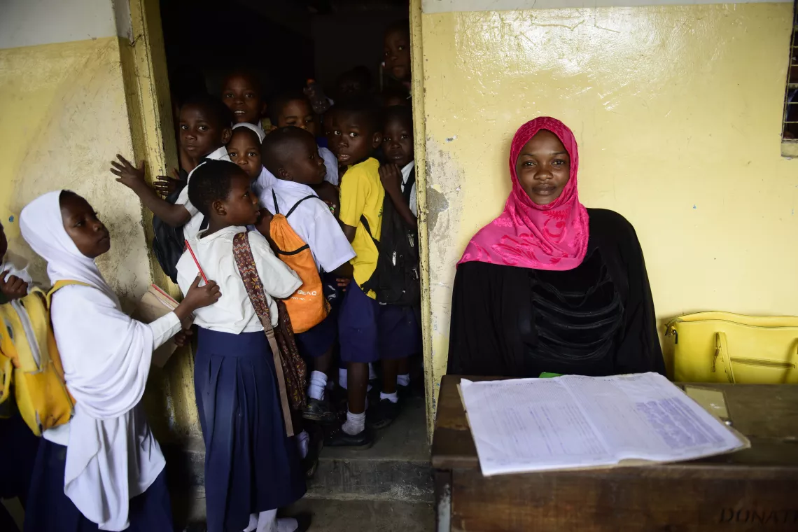 Teacher sitting by the door to the classroom, greeting her students. 