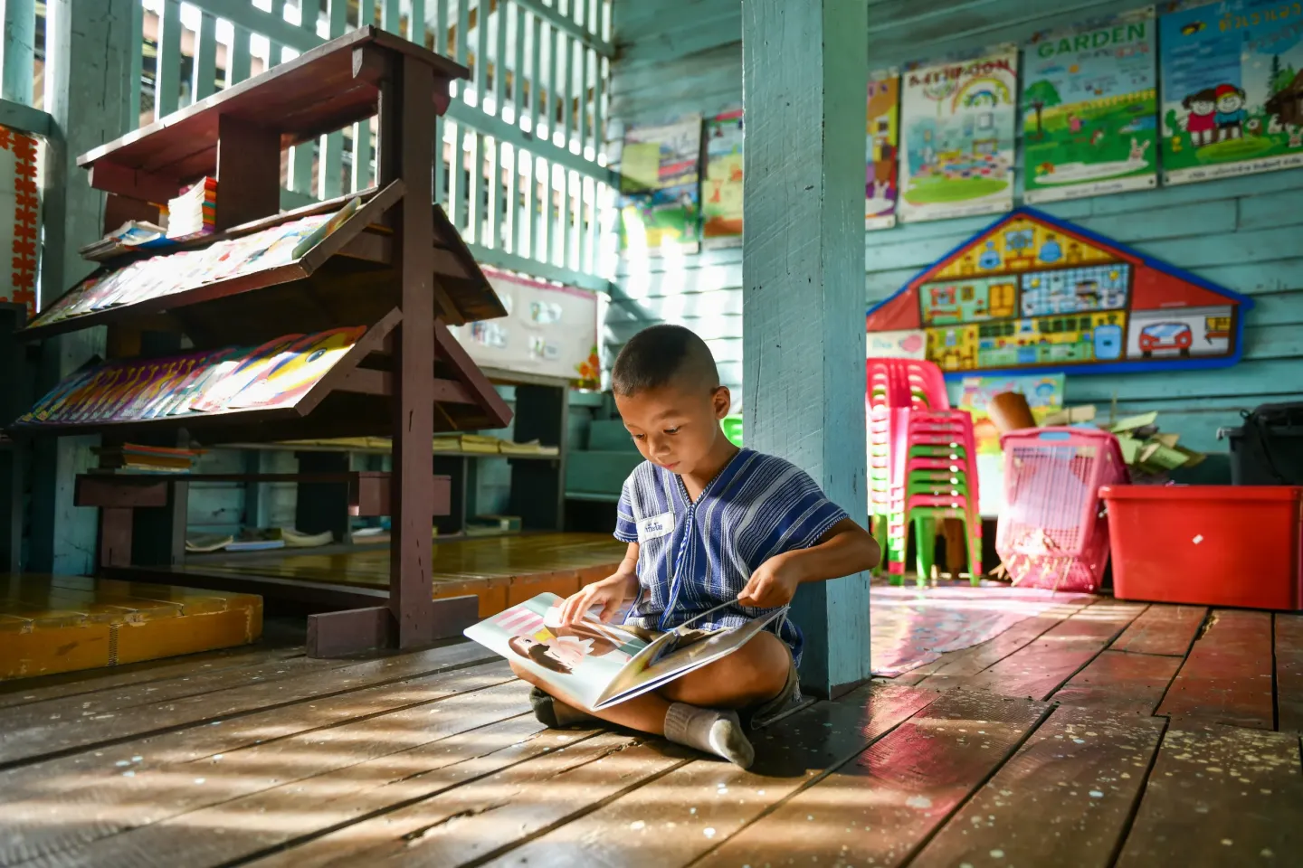 A young child sits on a wooden floor inside a simple, colorful classroom, quietly reading a picture book near a low bookshelf filled with learning materials.