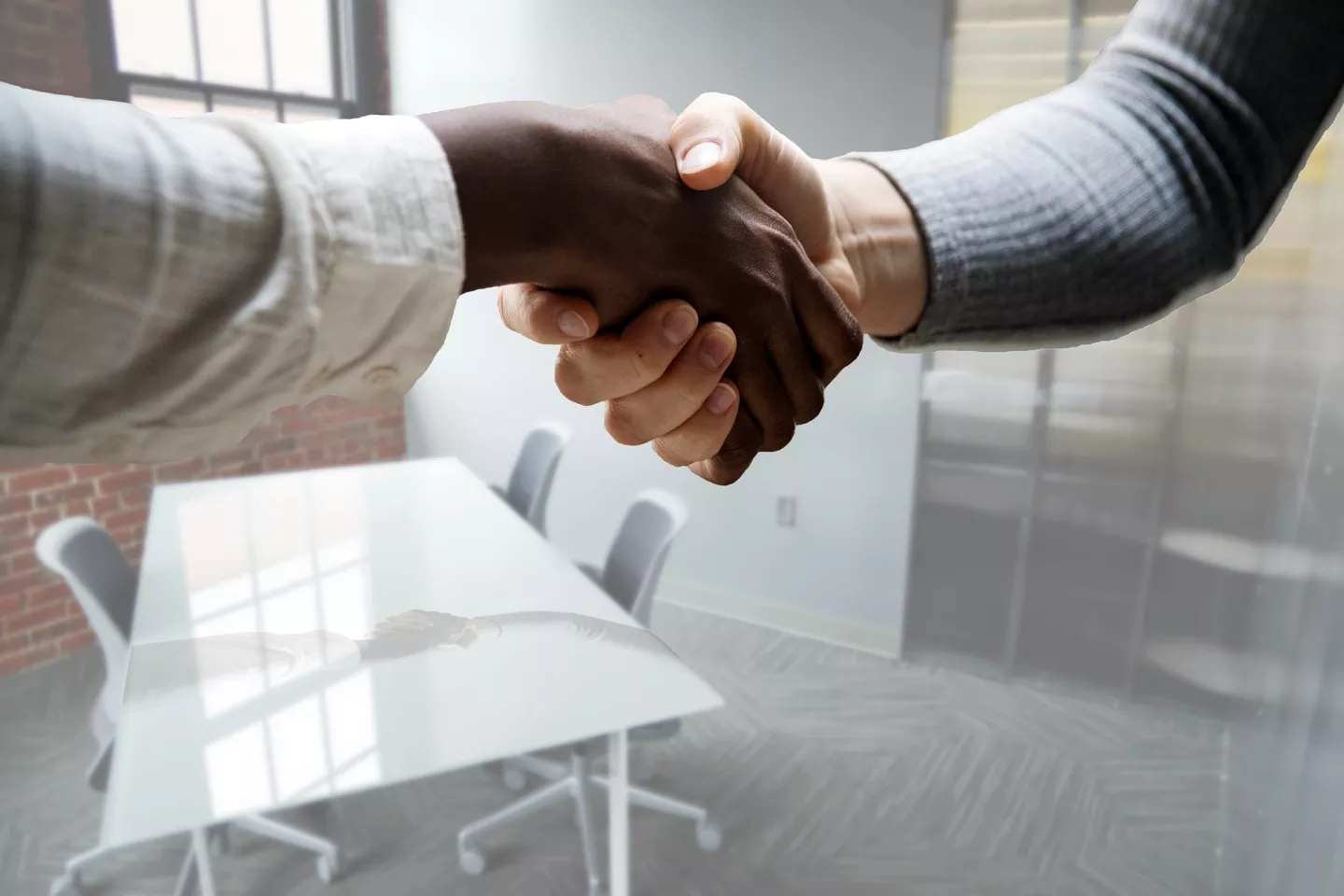 Handshaking. A hand on the left has dark skin, wearing a white shirt. The hand on the right has light skin, wearing a grey shirt.