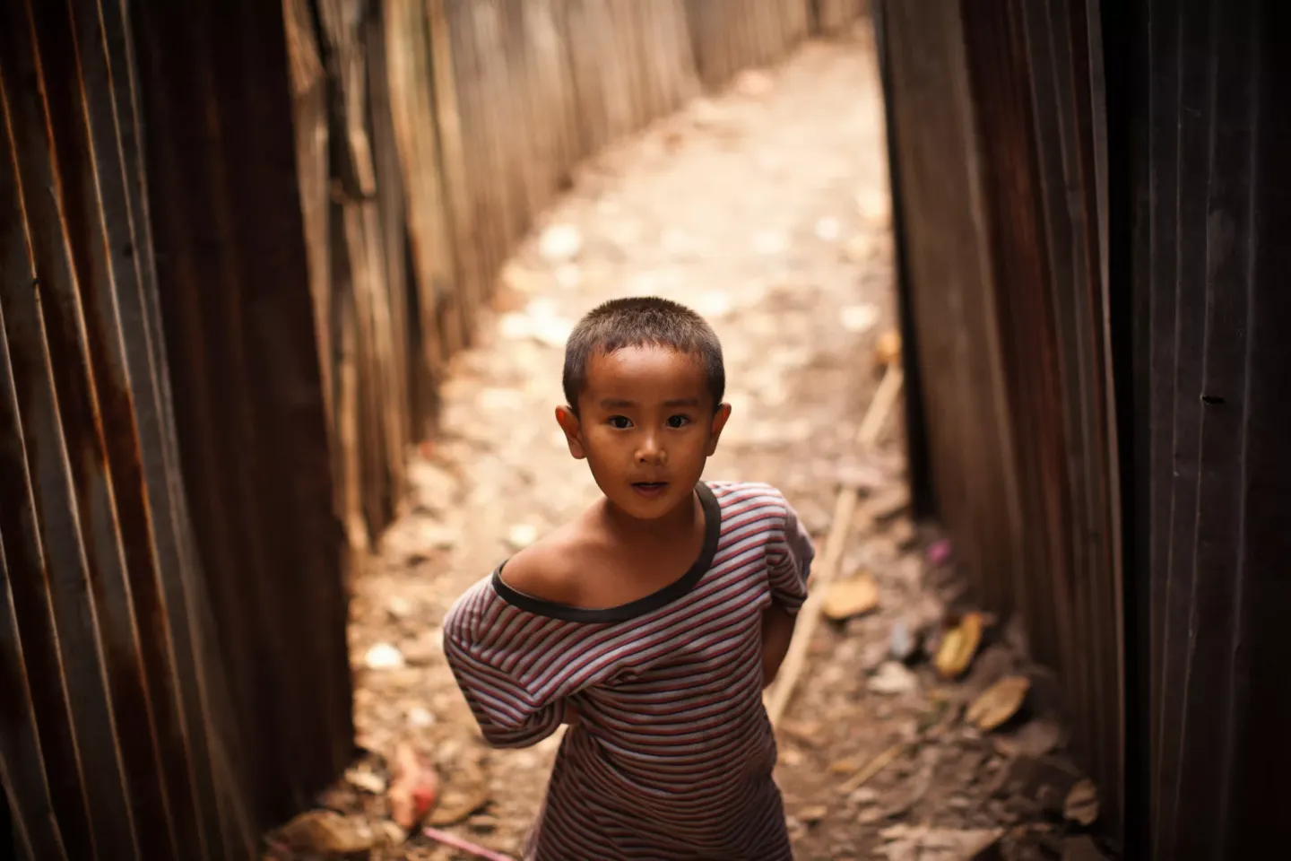 a young boy standing in a narrow alley between two corrugated metal walls. The boy has short hair and is wearing a striped shirt that is slipping off one shoulder. The alley is lined with dirt and debris, creating a rustic and rough environment. The boy looks directly at the camera with a curious and slightly surprised expression.