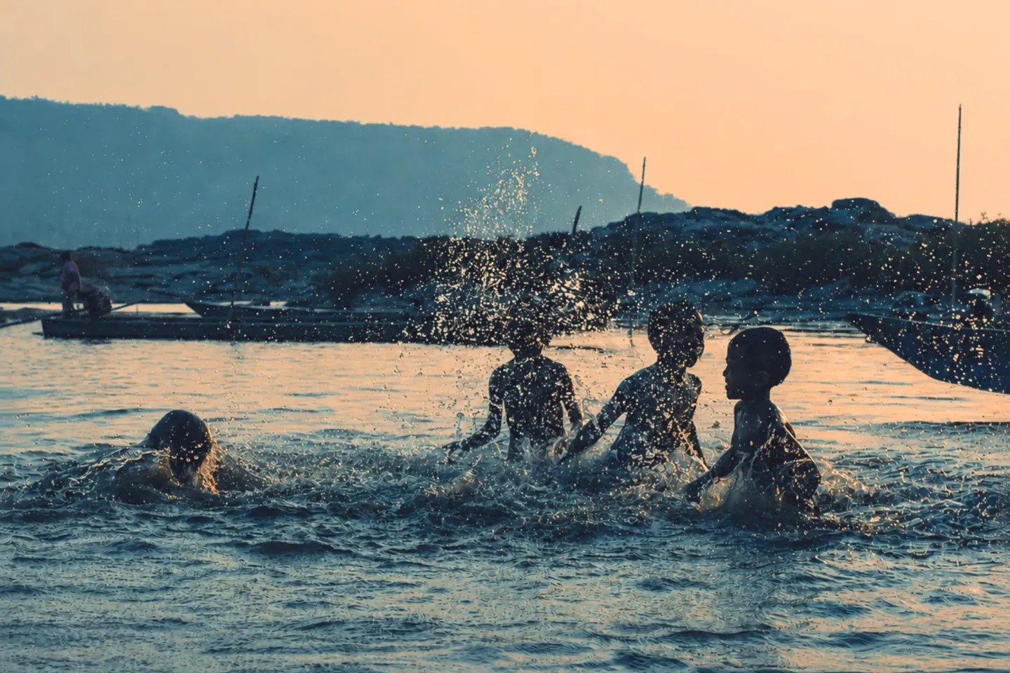 Several children splash and play in shallow water, with boats and a dark shoreline in the background.