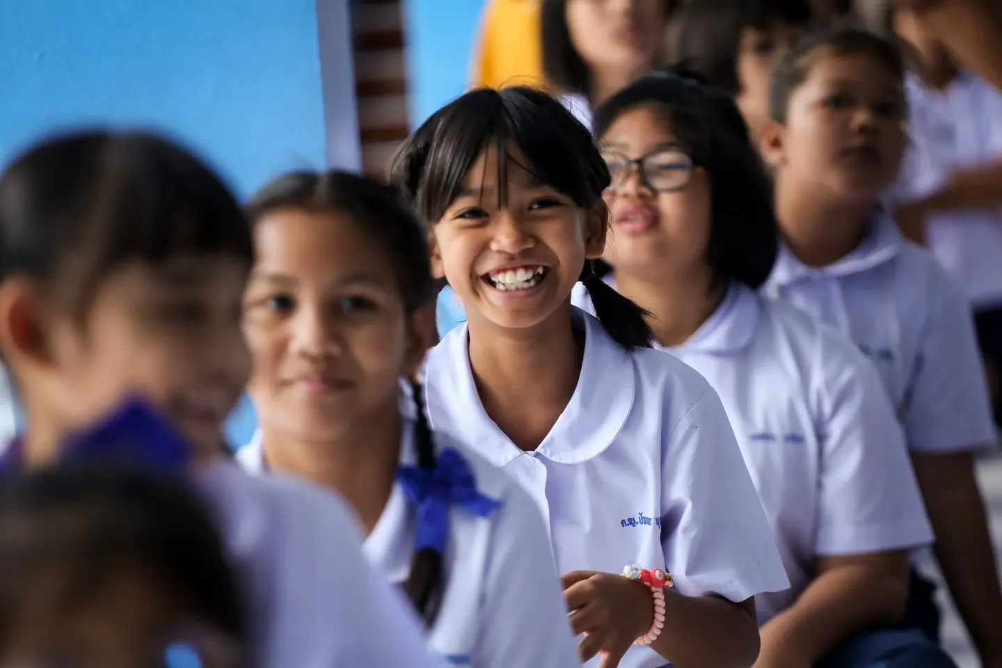 A smiling schoolgirl in a white uniform stands in line with other students, looking directly at the camera inside a school corridor.