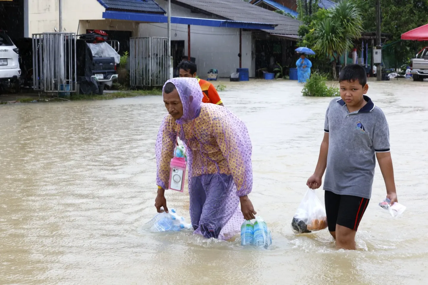 People wading through floodwater in the rain while carrying bags of drinking water and supplies.