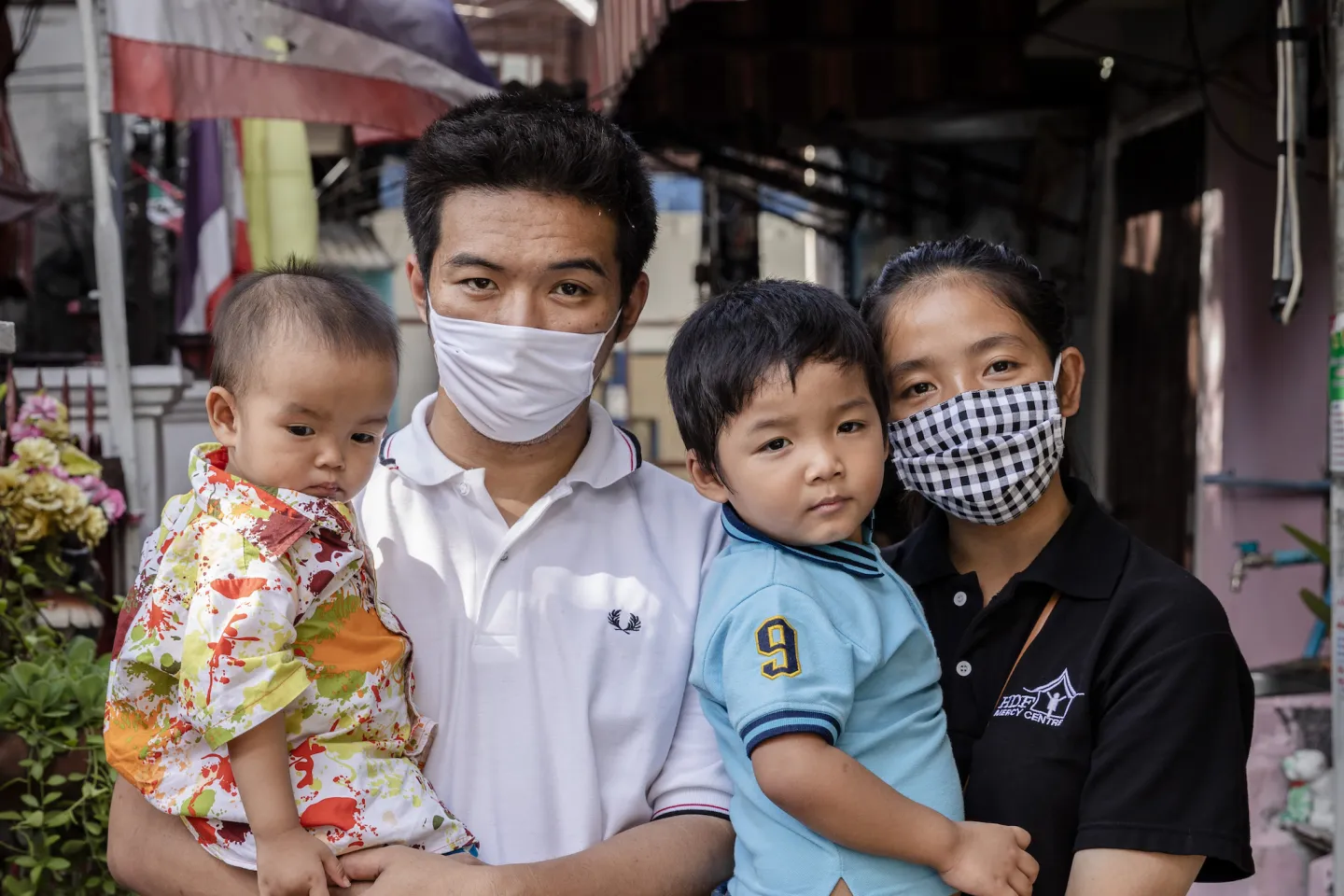 A family photograph. The father is wearing a face mask and is holding his youngest child. The mother, who is also wearing a mask, is holding her child in her arms. Neither of the children is wearing a mask.