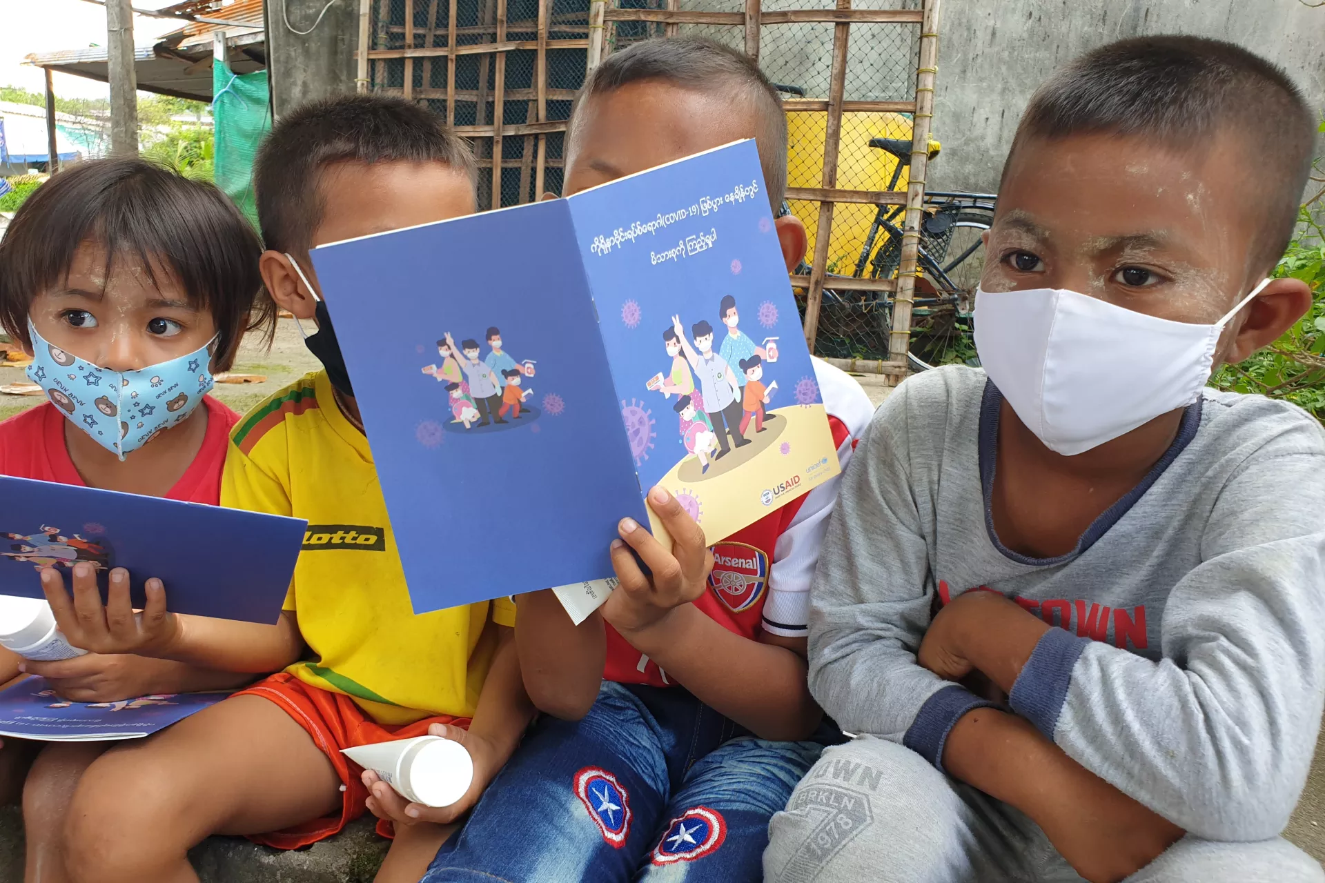 4 young children wearing a facial mask are sitting and reading a book.