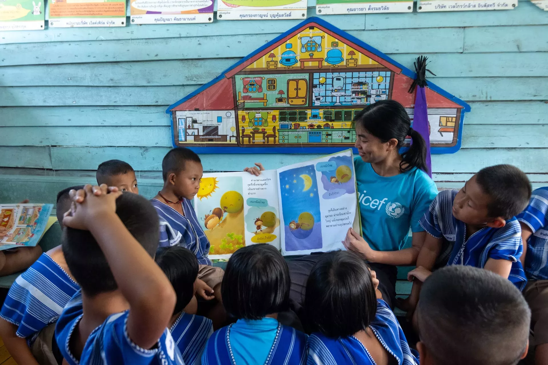 A woman in a UNICEF shirt reading a picture book to a group of children in blue uniforms sitting in a classroom with colorful educational posters.