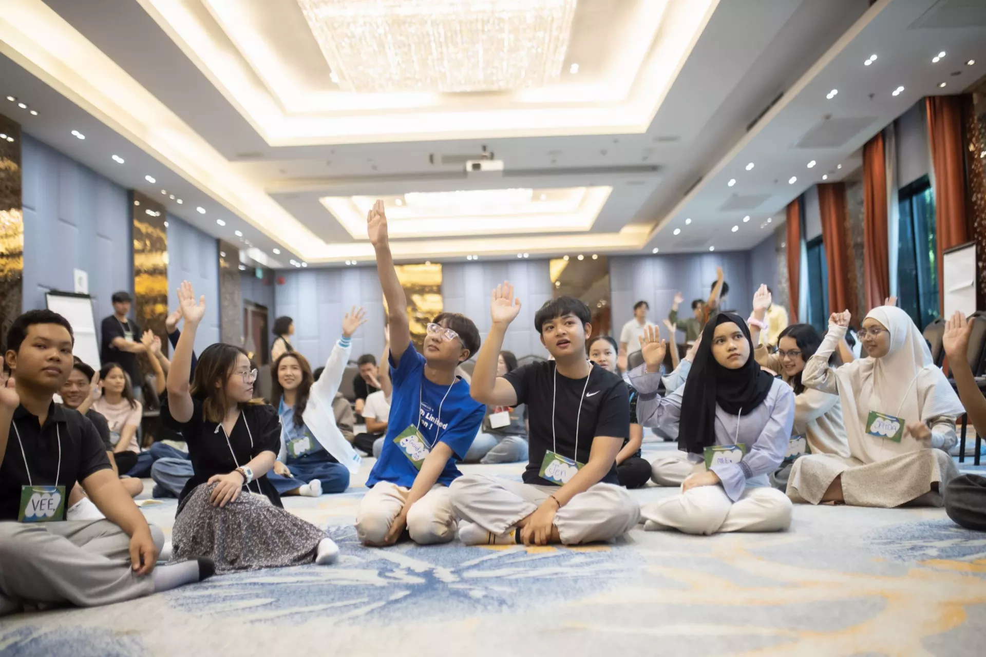 A group of diverse Thai youth sitting on the carpeted floor of a well-lit conference room, actively participating in a session. Many of them are raising their hands. The atmosphere is engaged and collaborative.