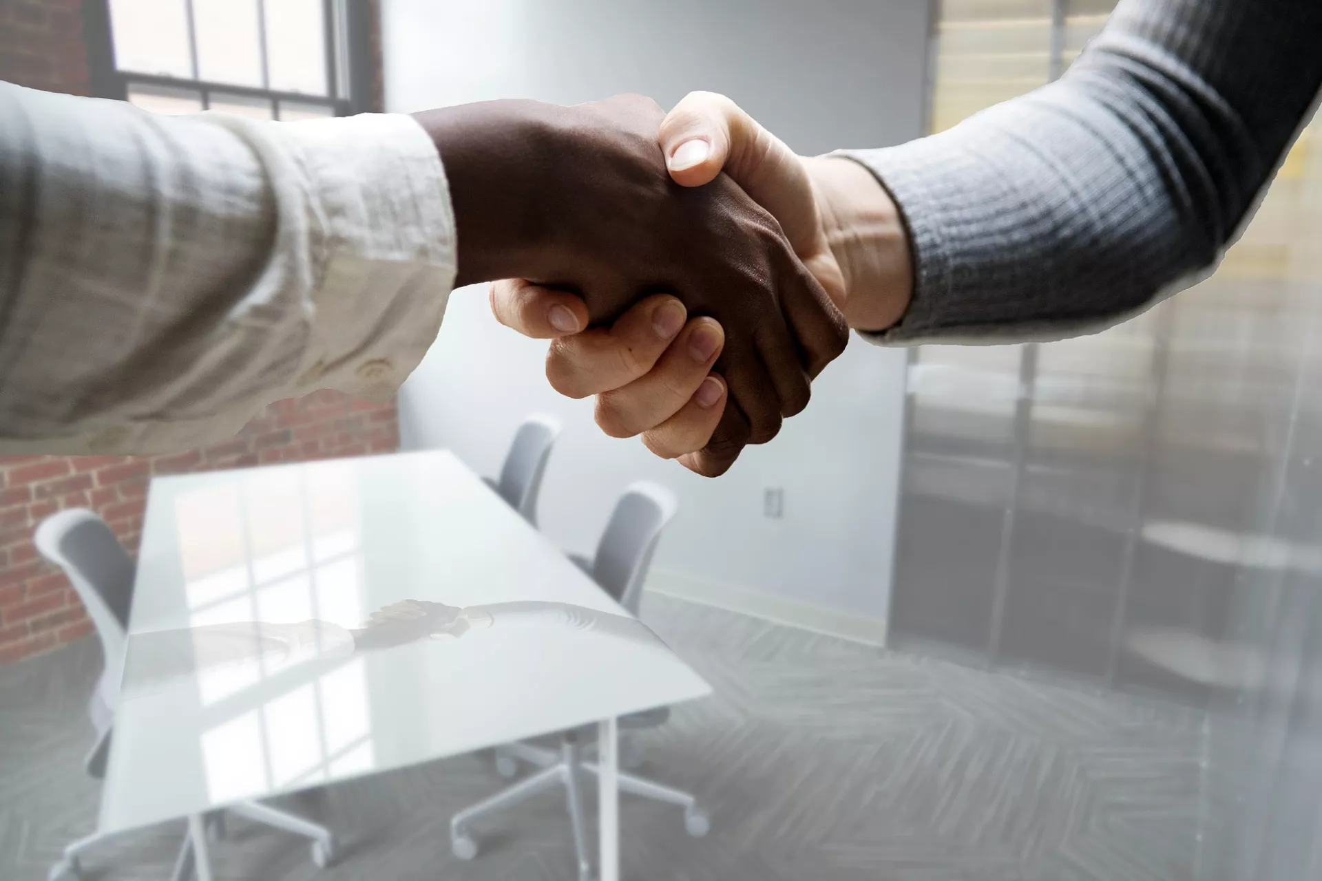 Handshaking. A hand on the left has dark skin, wearing a white shirt. The hand on the right has light skin, wearing a grey shirt.