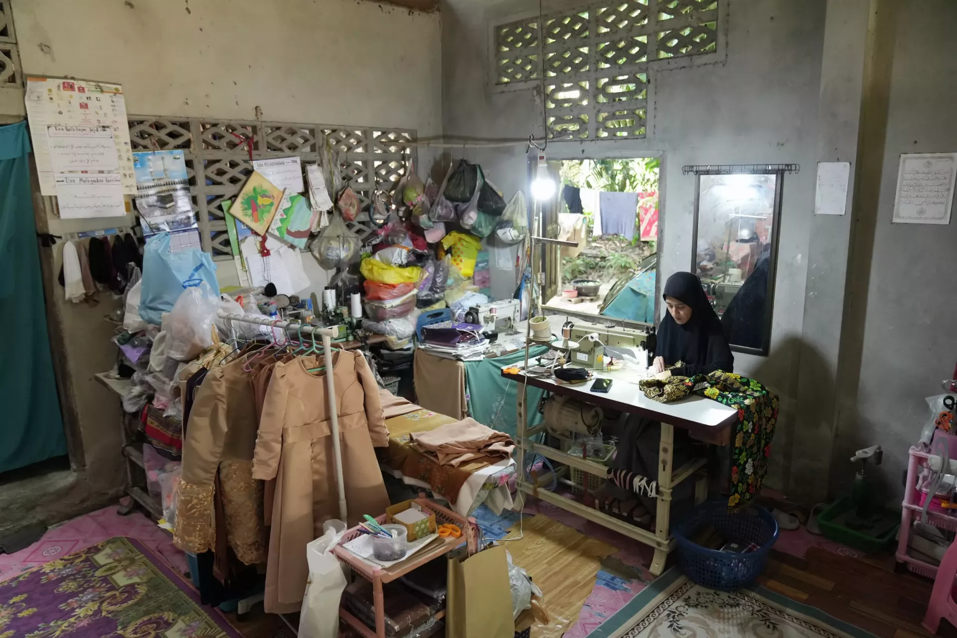 A woman in a hijab is focused on sewing colorful fabric at a sewing machine in a small, cluttered workshop filled with garments, tools, and supplies.