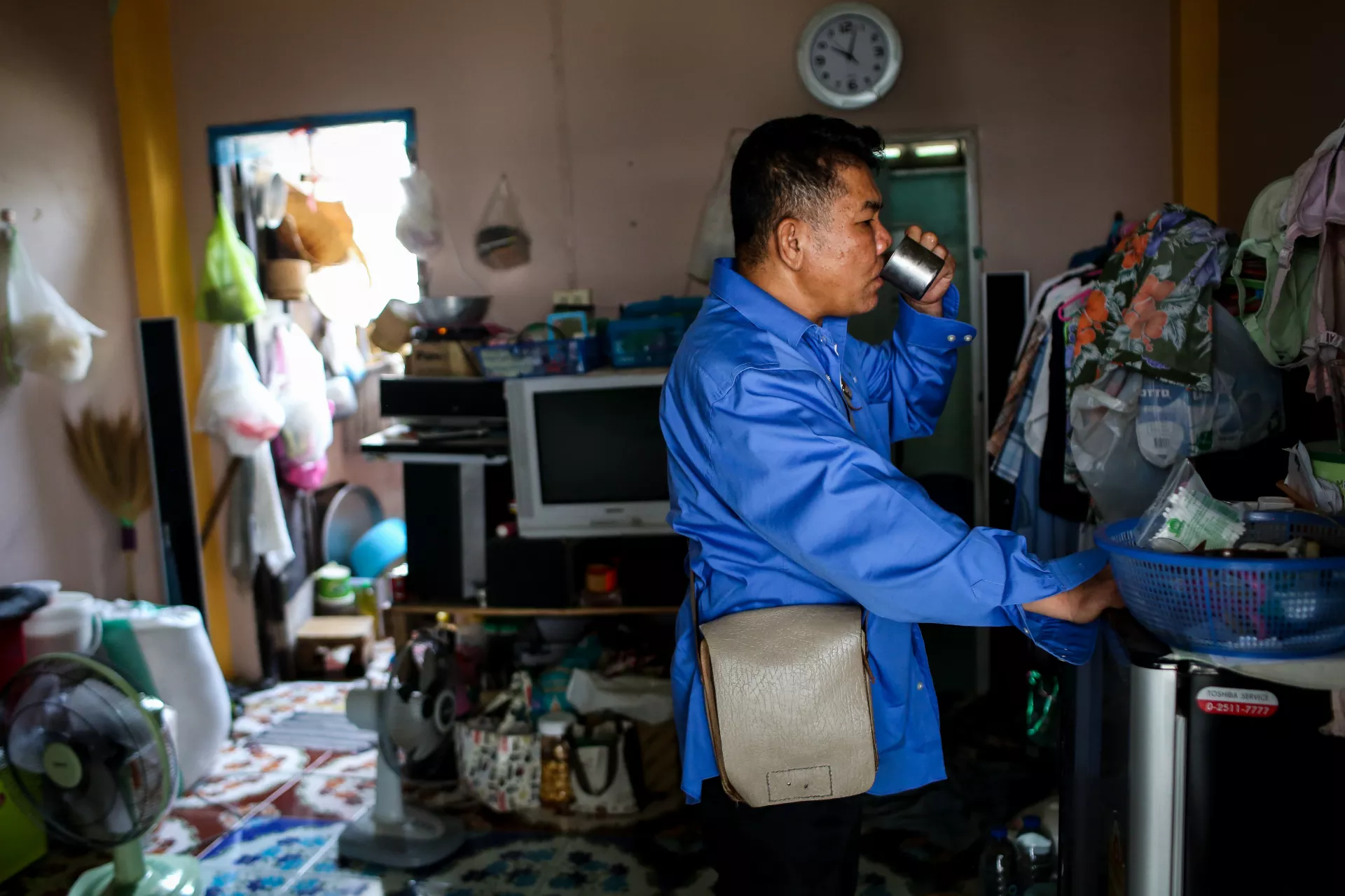A middle-age man taxi driver in a blue shirt is standing, drinking a cup of water in his house