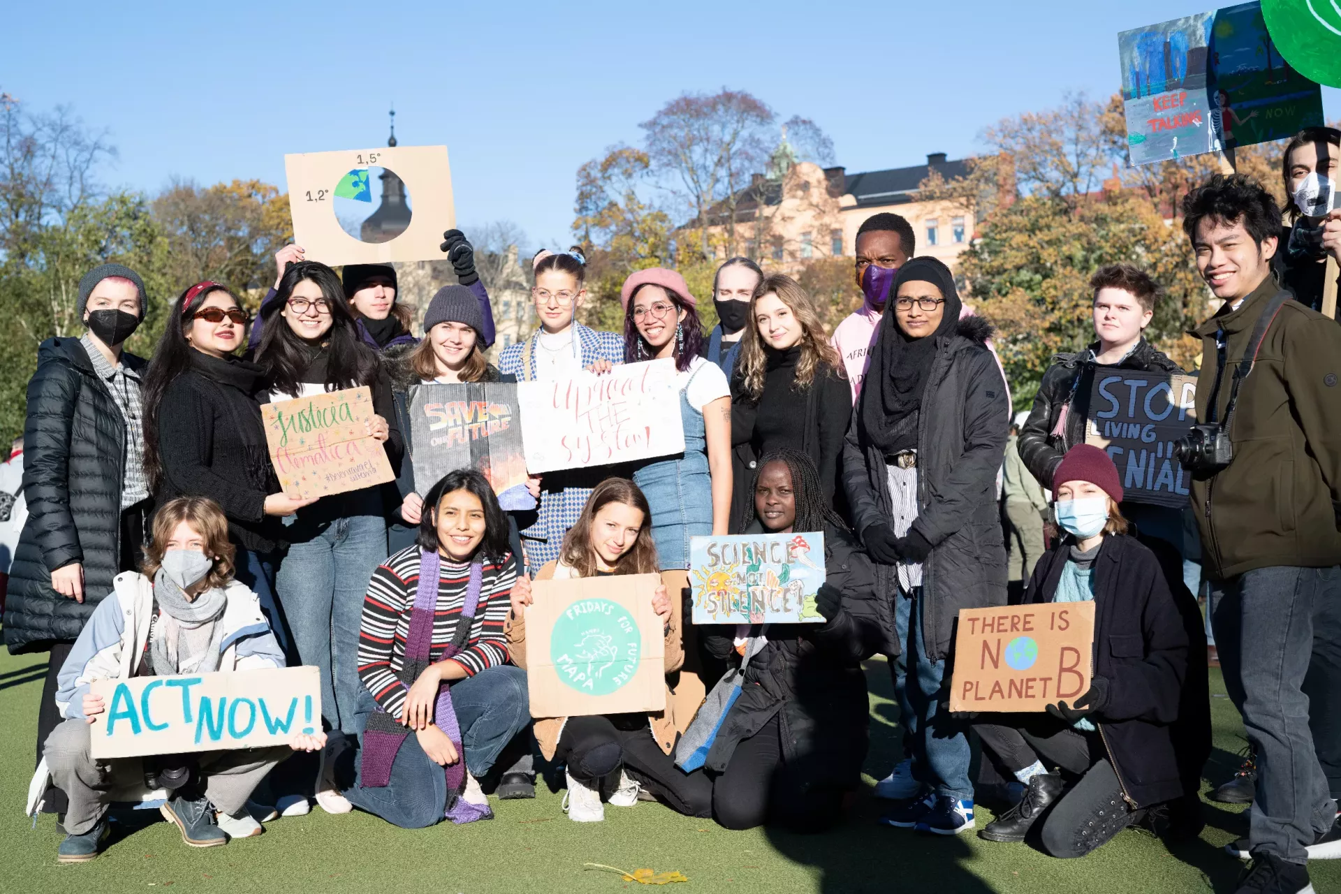 A group of diverse young people is gathered outdoors holding various signs advocating for climate action.