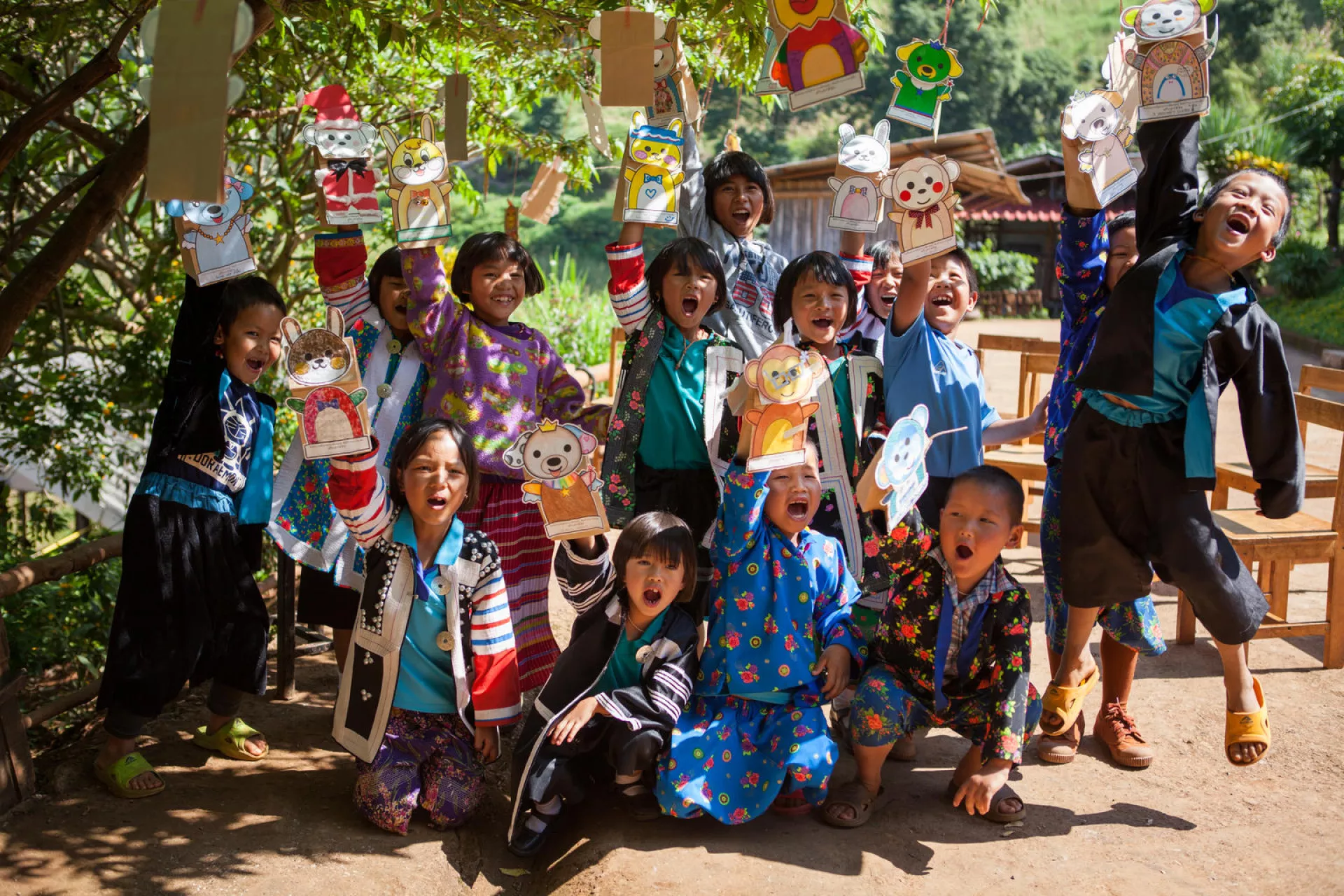 A group of hill-tribe children are raising their hands with paper hand puppets.