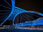 A modern pedestrian bridge with an arch and cables illuminated in blue light against the night sky. The structure's sleek design and vibrant lighting create a futuristic atmosphere, with high-rise buildings faintly visible in the background. A few people walk across the bridge, adding a sense of scale and activity to the scene.