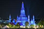 Phra Prang Wat Arun in Bangkok is illuminated in blue lights at night. The central prang and its smaller surrounding towers shine brightly against the dark sky. Trees and traditional Thai architecture are visible at the base, while faint city lights appear in the background.