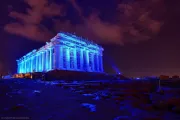 The Acropolis in Athens, Greece, illuminated with vibrant blue lights against a night sky. Scaffolding is present around parts of the ancient structure, suggesting ongoing preservation efforts. The dark sky with scattered clouds adds to the dramatic effect, creating a striking contrast with the rocky ground in the foreground.